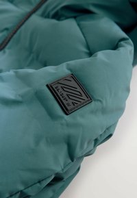 Türkise gepolsterte Jacke mit einem schwarzen quadratischen Logo-Patch, das abstrakte Linien und Koordinaten "86° 13' 49" W" auf dem Ärmel zeigt.