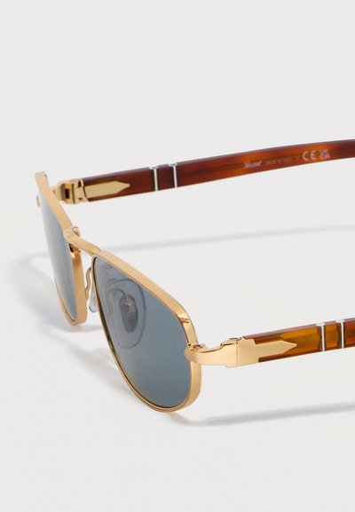 Γυαλιά ηλίου με χρυσαφί μεταλλικά πλαίσια, σκουρόγριους φακούς και καφέ βραχίονες με σχέδιο χελώνας, διακοσμημένους με χρυσαφί βέλη.