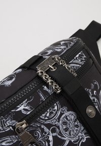 Black fabric bag featuring white ornate patterns, dual zippers, and chain-link hardware. Adjustable strap for secure fit.