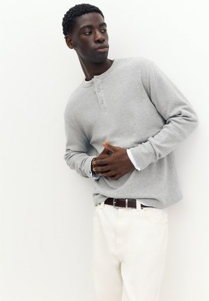Czarny mężczyzna ubrany w jasnoszary longsleeve typu henley i białe spodnie, patrzący na bok z rękami złożonymi z przodu.