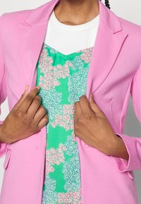 Różowa marynarka z klapkami, teksturowany materiał, noszona na zielonej sukience w kwiaty z różowymi akcentami. Ręce dostosowują przód marynarki.