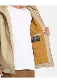 Giacca beige con cappuccio e chiusura a cerniera, con rivestimento interno giallo e una tasca con etichetta, indossata sopra una maglietta bianca.