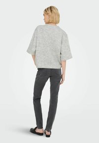 Femme blonde portant un pull court gris ample, un jean slim gris foncé et des mocassins noirs à enfiler, se tenant contre un fond uni.