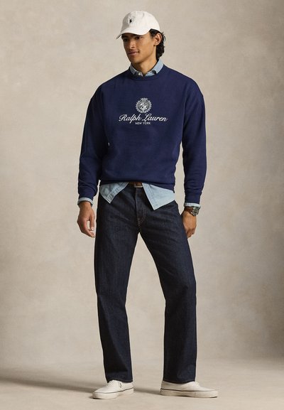 Μπλε ναυτικό φούτερ με λευκό λογότυπο, ανοιχτό μπλε πουκάμισο από κάτω, τζιν σκούρου χρώματος και λευκά αθλητικά παπούτσια, ολοκληρωμένο με λευκό καπέλο μπέιζμπολ.