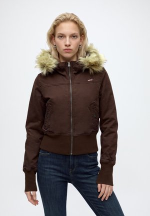 Jeune femme portant une veste marron zippée avec une bordure en fourrure sur la capuche et un jean bleu, debout sur un fond blanc uni.