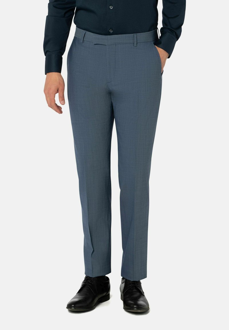 Man in slim-fit blauwe broek, zwarte leren schoenen en een donker knoopshirt met één hand in zijn zak.