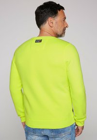 Sudadera amarilla neón con cuello redondo y puños acanalados. Cuenta con un parche del logo en la parte posterior y una tela suave y texturizada.