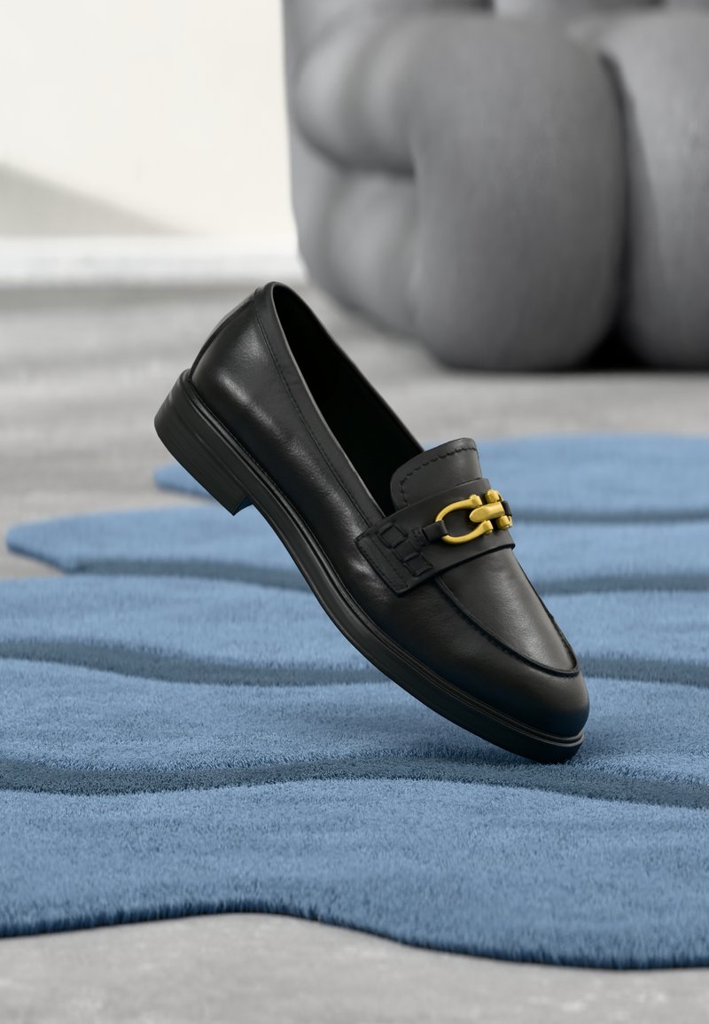 Mocassin en cuir noir avec un accent en chaîne dorée, semelle plate et détails de coutures, positionné au-dessus d'un tapis bleu texturé.