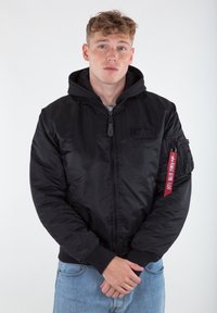 Black bomber jacket with a hood, zip closure, ribbed cuffs, and a patch on the chest. Features a red tag on the sleeve for detailing.
