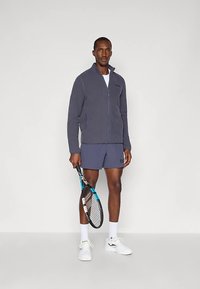 Muškarac u tamnoj jakni na zatvaranje, tamnoplavim šortsevima, bijelim tenisicama, drži reket za tenis, stoji naslonjen na jednostavnu svijetlu pozadinu.