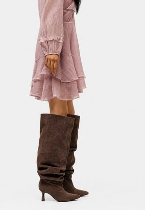 Femme portant une robe rose à couches et des bottes mousses en daim marron foncé montant jusqu'aux genoux avec des bouts pointus et des talons petits.