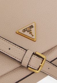 Beige texturovaná kabelka s trojúhelníkovým zlatým logem Guess a zlatým detailem s přezkou na odpovídajícím popruhu s kontrastním černým prošíváním.