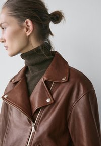 Giacca di pelle marrone con ampio colletto, elementi in metallo argentato e chiusura frontale con cerniera, indossata sopra un dolcevita verde scuro. Texture liscia.