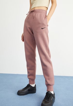 Personne portant un pantalon de jogging Nike mauve avec des baskets Nike noires et un top court beige, debout sur un sol bleu devant un mur blanc.