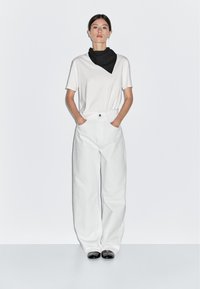 Bílý bavlněný trikoto s výstřihem do V, spárovaný s bílými širokými kalhotami. Černý čtvercový šátek nošený na krku; černé boty dotvářejí vzhled.