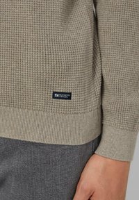 Jasno beżowy sweter o teksturowanej powierzchni z prążkowanymi mankietami i dolnym wykończeniem. Posiada małą granatową metkę z napisem "Elevated Casual" w pobliżu dolnej krawędzi.