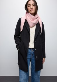 Cappotto di lana nero con rever a punta, chiusura con bottoni, abbinato a una sciarpa rosa texturizzata, maglione bianco a trecce e jeans a gamba larga blu.