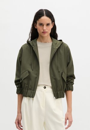 Vrouw met lang donker haar draagt een olijfgroene capuchontrui met rits over een beige shirt en hooggetailleerde crème broek, staand tegen een effen achtergrond.