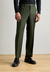 Pantalons moulants verts en tissu lisse, présentant un design à jambe droite, des plis avant et une fermeture à bouton unique. Chaussures noires à lacets.
