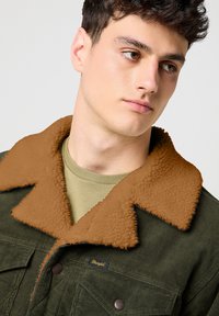 Chaqueta de pana verde oliva con un cuello de pelaje sintético marrón texturizado; cuenta con cierre de botones y bolsillos en el pecho. Camisa verde claro debajo.