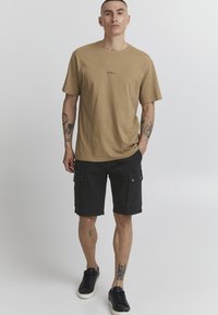 Beige katoenen t-shirt met een logo op de voorkant, gecombineerd met zwarte cargo shorts met zakken en zwarte sneakers. Eenvoudig ontwerp en comfortabele pasvorm.