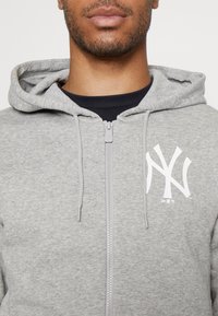 Sudadera gris con cremallera y bolsillo frontal, que presenta un logo blanco de los New York Yankees en el lado izquierdo del pecho, hecha de una tela suave y texturizada.