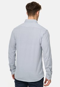 Das Langarmhemd hat einen weißen Grundton mit dunkelblauen geometrischen Mustern, einen Kentkragen, Knopfmanschetten und einen abgerundeten Saum.