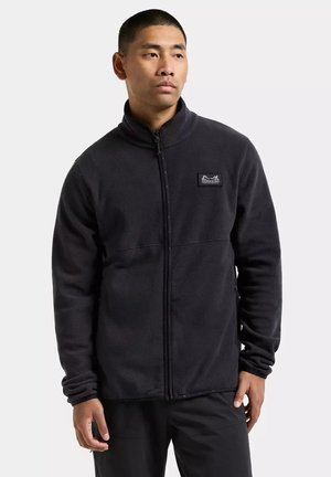 Man draagt een zwarte fleecejas met rits en een hoge kraag, met een logopatch op de linkerborst, staand tegen een effen achtergrond.