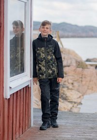 Vattentät kamouflagejacka med svarta ärmar, i kombination med svarta byxor. Funktioner inkluderar dragkedjor och en huva. Miljö: stenig brygga.