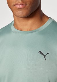Camiseta deportiva en teal claro, fabricada con un tejido suave, que presenta un logo negro de Puma en el pecho y un diseño clásico de cuello redondo.