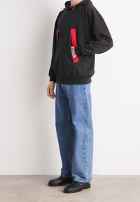 Felpa nera con cappuccio e dettagli rossi, con due tasche frontali, abbinata a jeans blu larghi e scarpe nere con dettagli in rilievo.