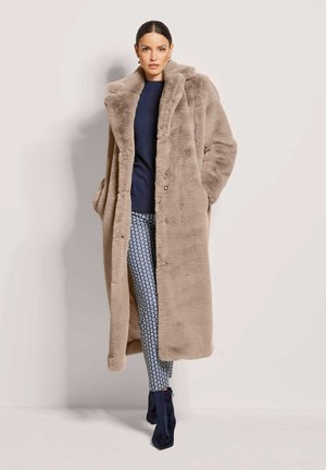 Lange, beige jas van nepbont met een brede kraag, knoopsluiting en voorzakken, gecombineerd met blauwe leggings met patroon en donkere enkellaarsjes.