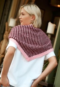 Châle tricoté avec une bordure rose, présentant un motif géométrique en marron et rose. Porté drapé sur les épaules avec un t-shirt blanc en dessous.