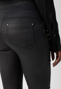 Gros plan d'une personne portant un pantalon noir ajusté avec des détails sur les poches arrière et une chemise noire à manches longues avec des poignets boutonnés.