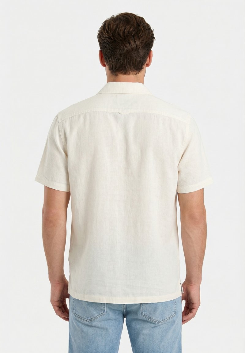 Homme aux cheveux courts et bruns portant une chemise en lin crème à manches courtes et un jean bleu clair, tourné dos à l'appareil photo.