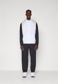 Tracksuit med en hvid og sort farveskema, med en lynlåslukning jakke og bukser med elastisk talje. Lavet af let, åndbart materiale.