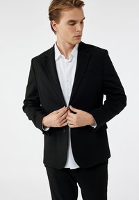 Schwarzer Blazer mit einem einzigen Knopfverschluss, ausgestattet mit Reverskragen und einer Brusttasche. Getragen über einem weißen Hemd, mit glatter Stofftextur.