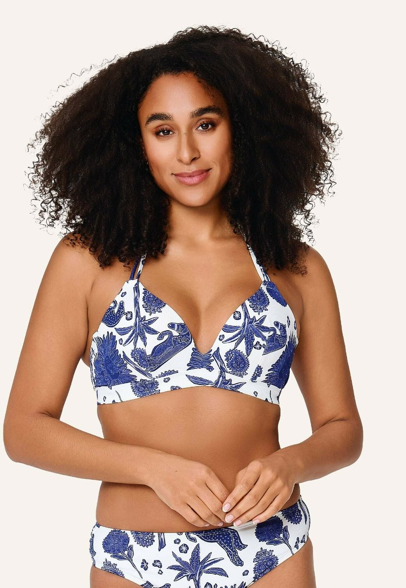 Mujer con cabello rizado que lleva un bikini blanco a juego con estampado botánico y de animales en azul, de pie con las manos suavemente entrelazadas.