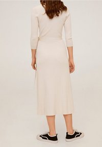 Femme aux cheveux bruns ondulés portant une robe midi crème à manches longues et des baskets noires et blanches, debout de dos face au spectateur.