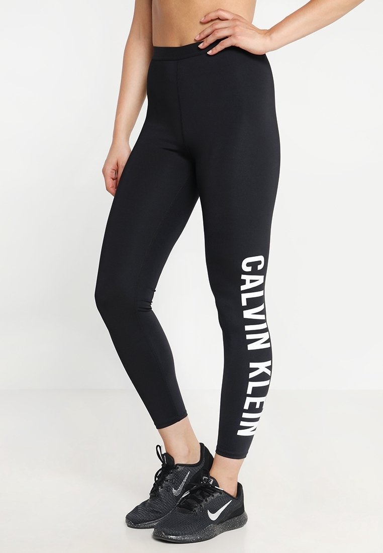 Czarne legginsy z wysokim stanem, z białym logo "CALVIN KLEIN" biegnącym pionowo wzdłuż lewego nogawki. Gładki, elastyczny materiał.