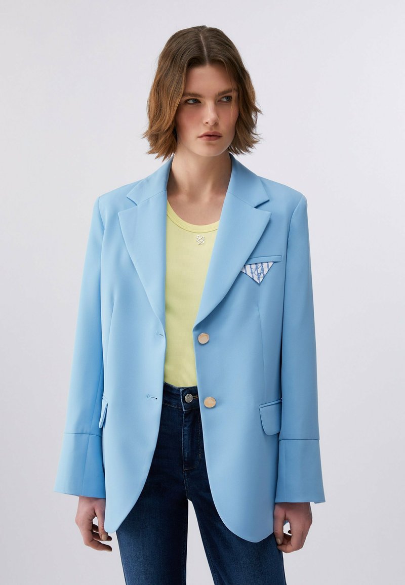Jeune femme aux cheveux bruns courts porte un blazer bleu clair, un haut jaune, un jean foncé et un mouchoir de poche plié à motifs sur fond uni.