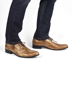 Gouden leren schoenen met een slangenhuidpatroon, puntige neus, zwarte vetersluiting, gecombineerd met donkere denim jeans.