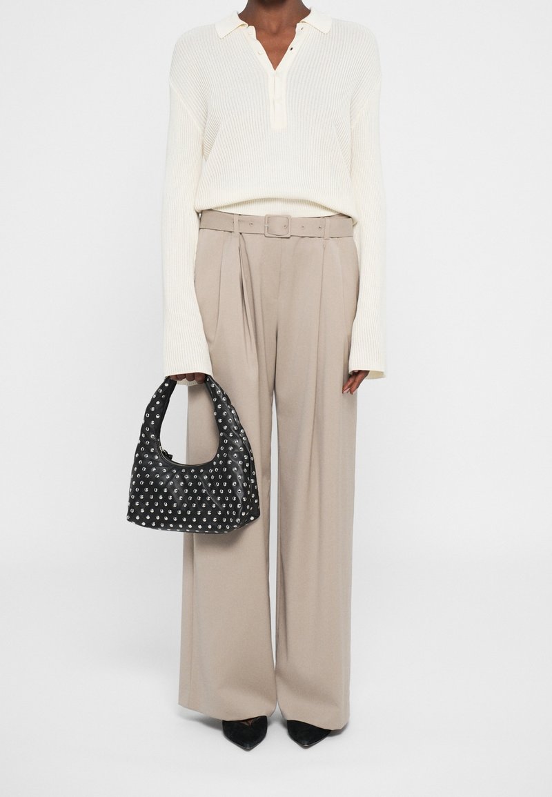 Personne portant un pull col blanc, un pantalon large beige avec ceinture, des chaussures noires pointues, tenant un sac à main noir à clous.