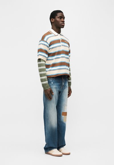 Νέος άνδρας που στέκεται με τζιν με μπλε μπαλώματα, πολλαπλές ριγέ μπλούζες σε καφέ, μπλε, πράσινο και λευκό, και μπεζ αθλητικά παπούτσια, μπροστά από ένα απλό φόντο.