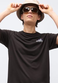 Νεαρό άτομο που φοράει μπεζ καπέλο τύπου κάδο, σκούρα γυαλιά ηλίου και μαύρο μπλουζάκι Patagonia με λογότυπο βουνού και το κείμενο «Save Our Home Planet».