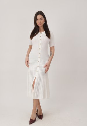 Vrouw met lang donker haar, gekleed in een witte geribbelde jurk met knopen en een voorsplit, en bordeauxrode hoge hakken, staand tegen een effen achtergrond.