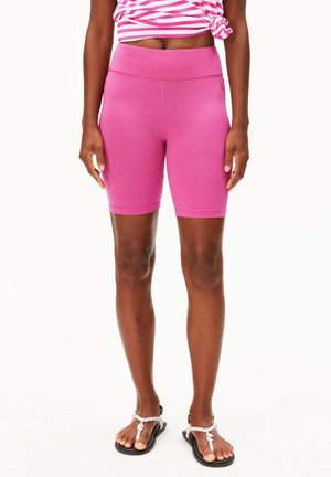 Frau trägt hoch taillierte rosa Radlerhosen, ein gestreiftes pink-weißes, geknotetes Oberteil und schwarze Sandalen mit weißen Seilen, steht vor weißem Hintergrund.