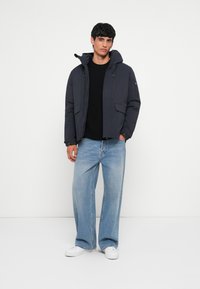 Mørkeblå pufferjakke med hætte og frontlommer, parret med lyseblå bredbenede jeans og hvide sneakers. Simpel, funktionel design.