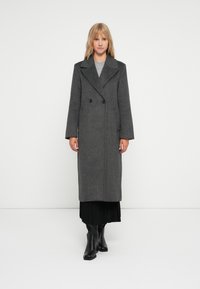 Abrigo de lana gris con un diseño de doble botonadura, grandes solapas y dos botones negros. Tiene mangas largas y llega hasta la mitad de la pantorrilla.