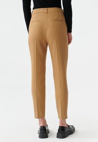 Pantalon beige sur mesure avec une texture lisse, coupe ajustée et deux poches arrière. Porté avec des chaussures à plateforme noires, vu de dos.